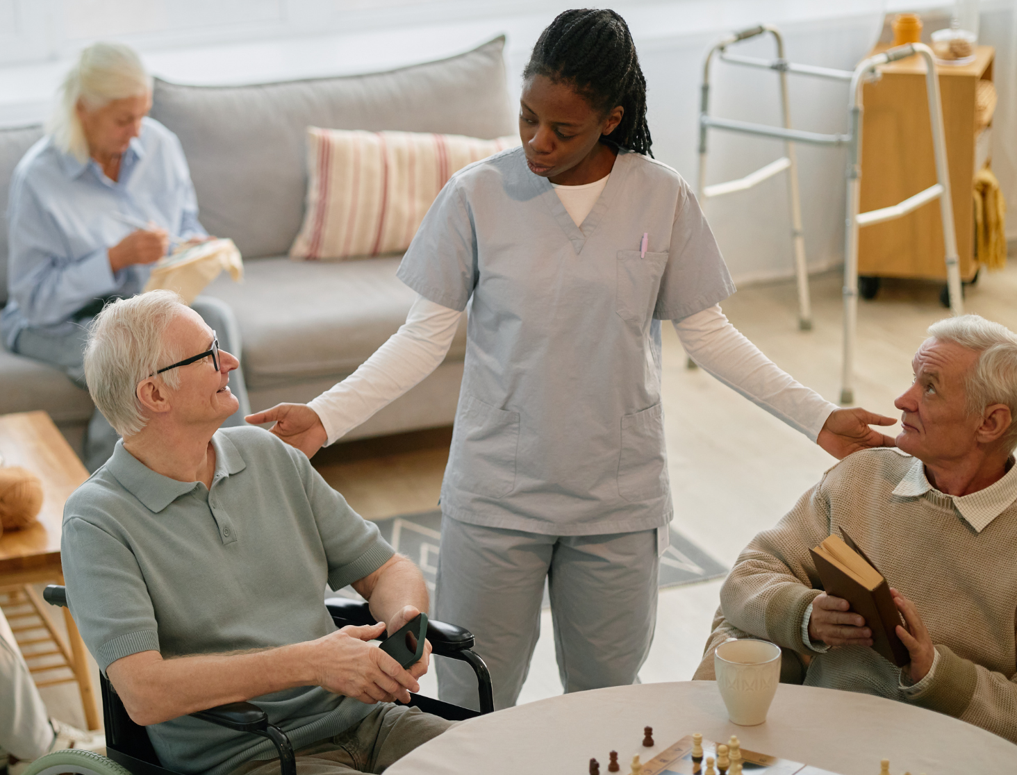 senior aid talking to two senior men at an assisted living facility, with two seniors behind them knitting